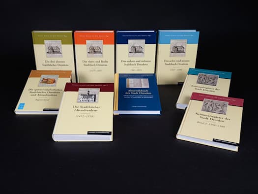 Acht dicke, beige Hardcover-Bücher mit farbigen Kopfleisten in Grün, Orange, Blau und Rot sind in zwei Reihen auf schwarzem Hintergrund aufgestellt. Auf den Einbänden sind historische Grafiken und Titel wie „Kriminalregister der Stadt Dresden“ zu sehen.
