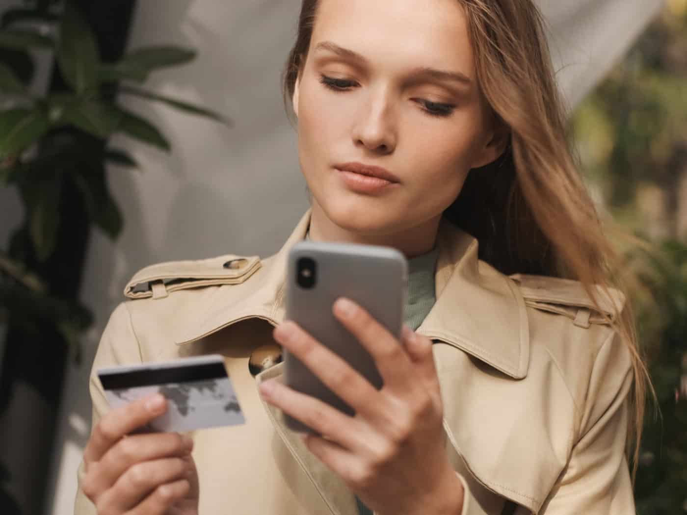 Eine junge Frau mit langen blonden Haaren im beigen Mantel schaut konzentriert auf ihr Smartphone. In der anderen Hand hält sie eine Plastikkarte. Im Hintergrund sind unscharf grüne Pflanzen im Freien zu sehen.