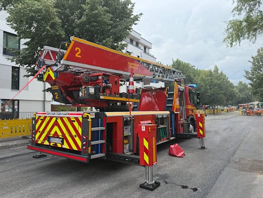 Ein rotes Feuerwehr-Drehleiterfahrzeug steht mit ausgefahrenen Stützen auf einer gesperrten Straße in Dresden. Im Hintergrund sind ein modernes Wohnhaus, grüne Bäume und eine Baustelle zu sehen.