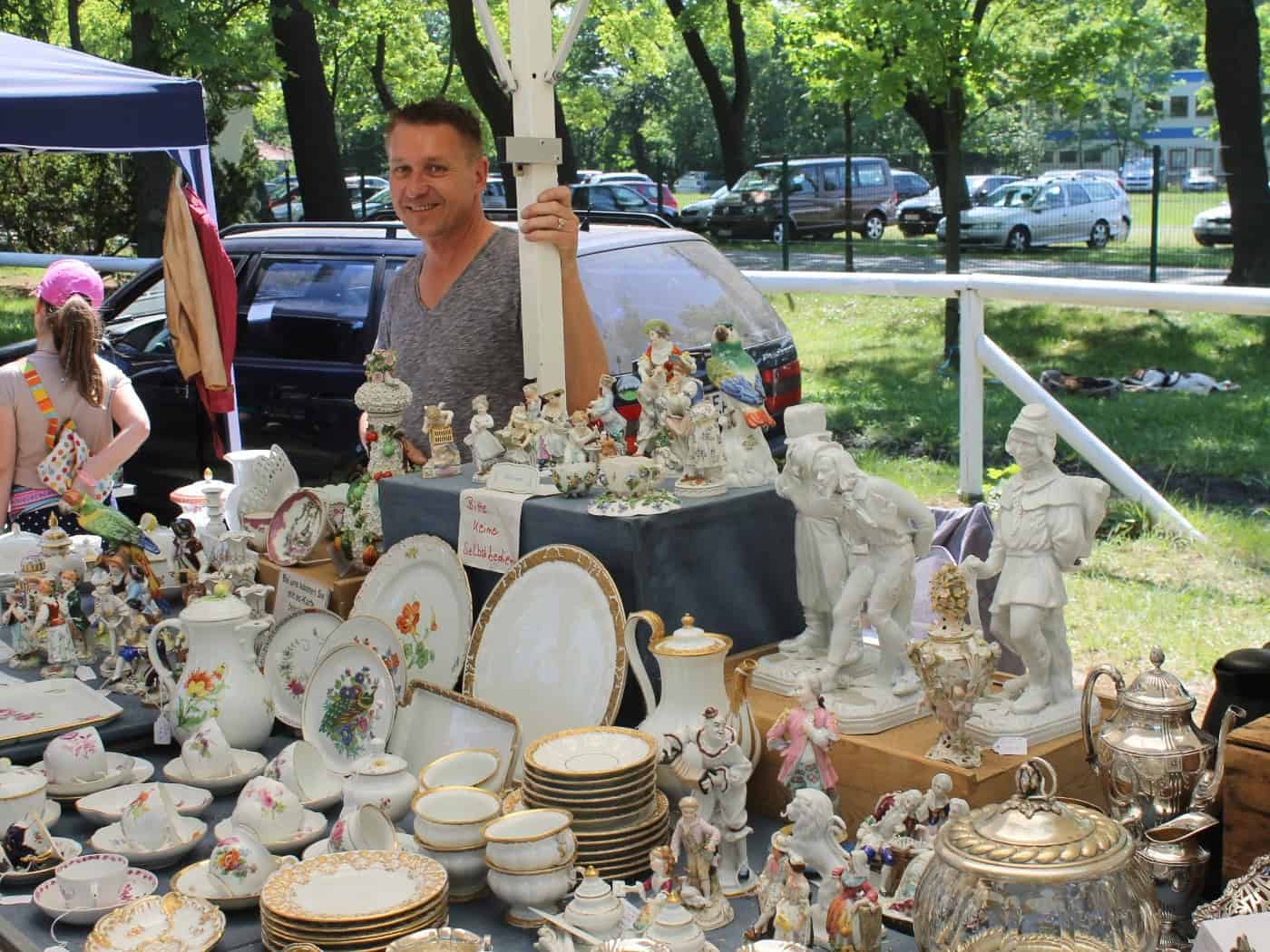Ein lächelnder Händler an seinem Flohmarktstand voller antiker Schätze. Auf dem Tisch stehen filigrane Porzellanfiguren, reich verzierte Kaffeekannen und Sammeltassen mit bunten Blumenmustern und Goldrand. Im Hintergrund Parkplätze und grüne Bäume.