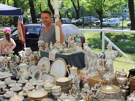Ein lächelnder Händler an seinem Flohmarktstand voller antiker Schätze. Auf dem Tisch stehen filigrane Porzellanfiguren, reich verzierte Kaffeekannen und Sammeltassen mit bunten Blumenmustern und Goldrand. Im Hintergrund Parkplätze und grüne Bäume.