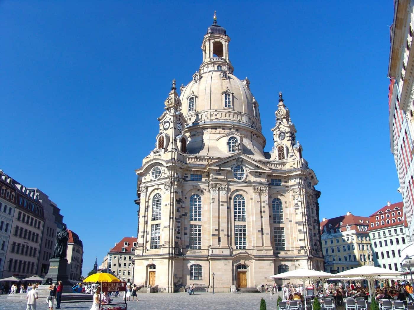 Die barocke Frauenkirche in Dresden fotografiert von unten gegen einen klaren blauen Himmel. Auf dem gepflasterten Neumarkt davor stehen weiße Sonnenschirme von Cafés und Passanten spazieren umher. Links im Vordergrund ist das dunkle Lutherdenkmal zu sehen.