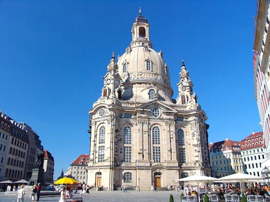 Die barocke Frauenkirche in Dresden fotografiert von unten gegen einen klaren blauen Himmel. Auf dem gepflasterten Neumarkt davor stehen weiße Sonnenschirme von Cafés und Passanten spazieren umher. Links im Vordergrund ist das dunkle Lutherdenkmal zu sehen.