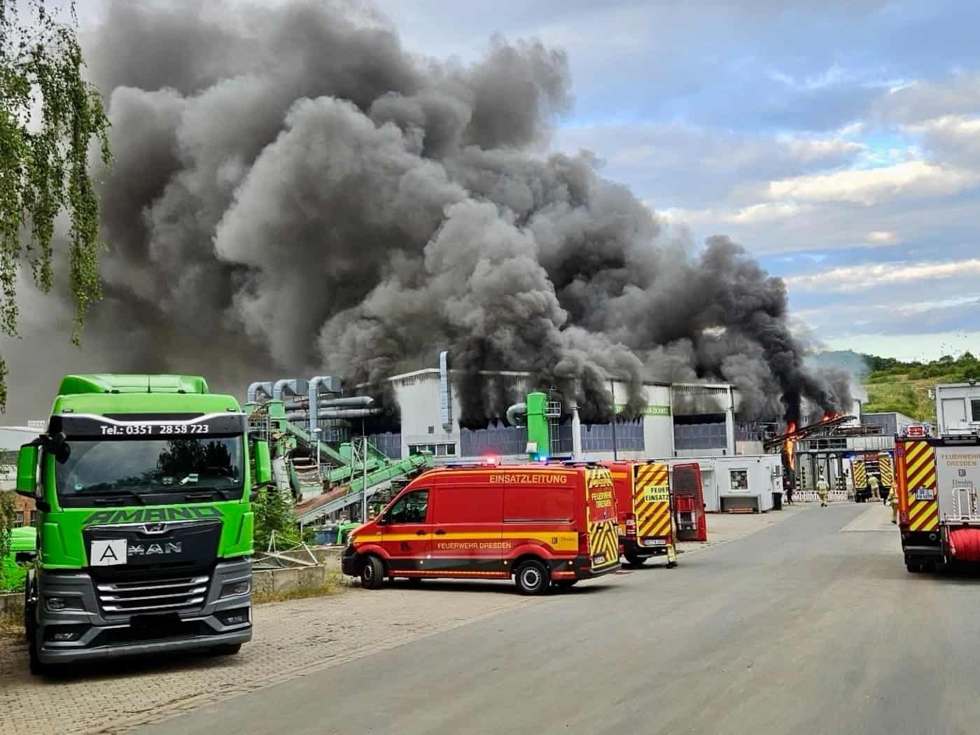 Feuerwehrfahrzeuge stehen vor einer Industriehalle in Dresden-Lockwitz, aus der eine riesige, dichte schwarze Rauchsäule in den bewölkten Himmel steigt. Links parkt ein grüner Lkw, im Hintergrund sind Einsatzkräfte im Einsatz.