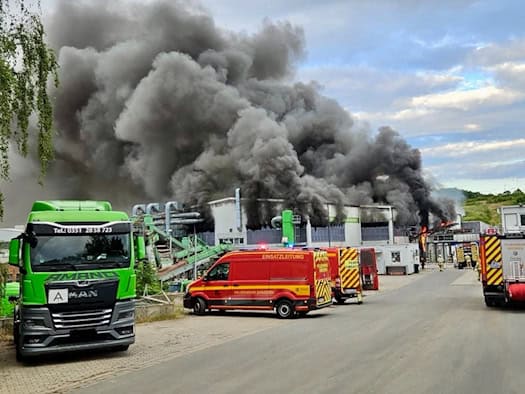 Feuerwehrfahrzeuge stehen vor einer Industriehalle in Dresden-Lockwitz, aus der eine riesige, dichte schwarze Rauchsäule in den bewölkten Himmel steigt. Links parkt ein grüner Lkw, im Hintergrund sind Einsatzkräfte im Einsatz.