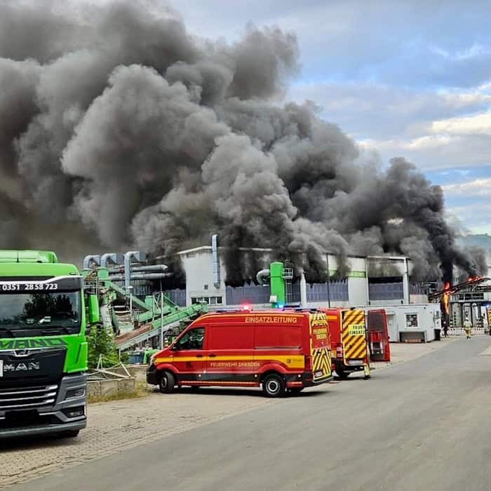 Feuerwehrfahrzeuge stehen vor einer Industriehalle in Dresden-Lockwitz, aus der eine riesige, dichte schwarze Rauchsäule in den bewölkten Himmel steigt. Links parkt ein grüner Lkw, im Hintergrund sind Einsatzkräfte im Einsatz.