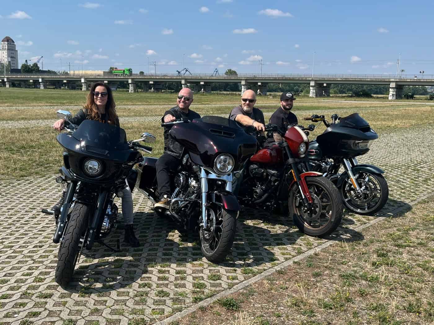 Eine Frau und drei Männer sitzen von links nach rechts lächelnd auf vier großen, dunklen Motorrädern der Marke Harley-Davidson. Sie stehen auf Rasengittersteinen einer weiten Wiese vor einer langen Betonbrücke im Dresdner Ostragehege.