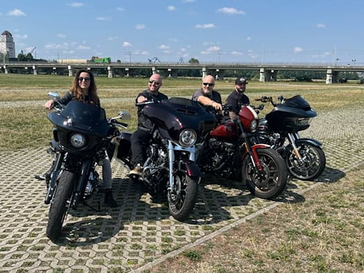 Eine Frau und drei Männer sitzen von links nach rechts lächelnd auf vier großen, dunklen Motorrädern der Marke Harley-Davidson. Sie stehen auf Rasengittersteinen einer weiten Wiese vor einer langen Betonbrücke im Dresdner Ostragehege.