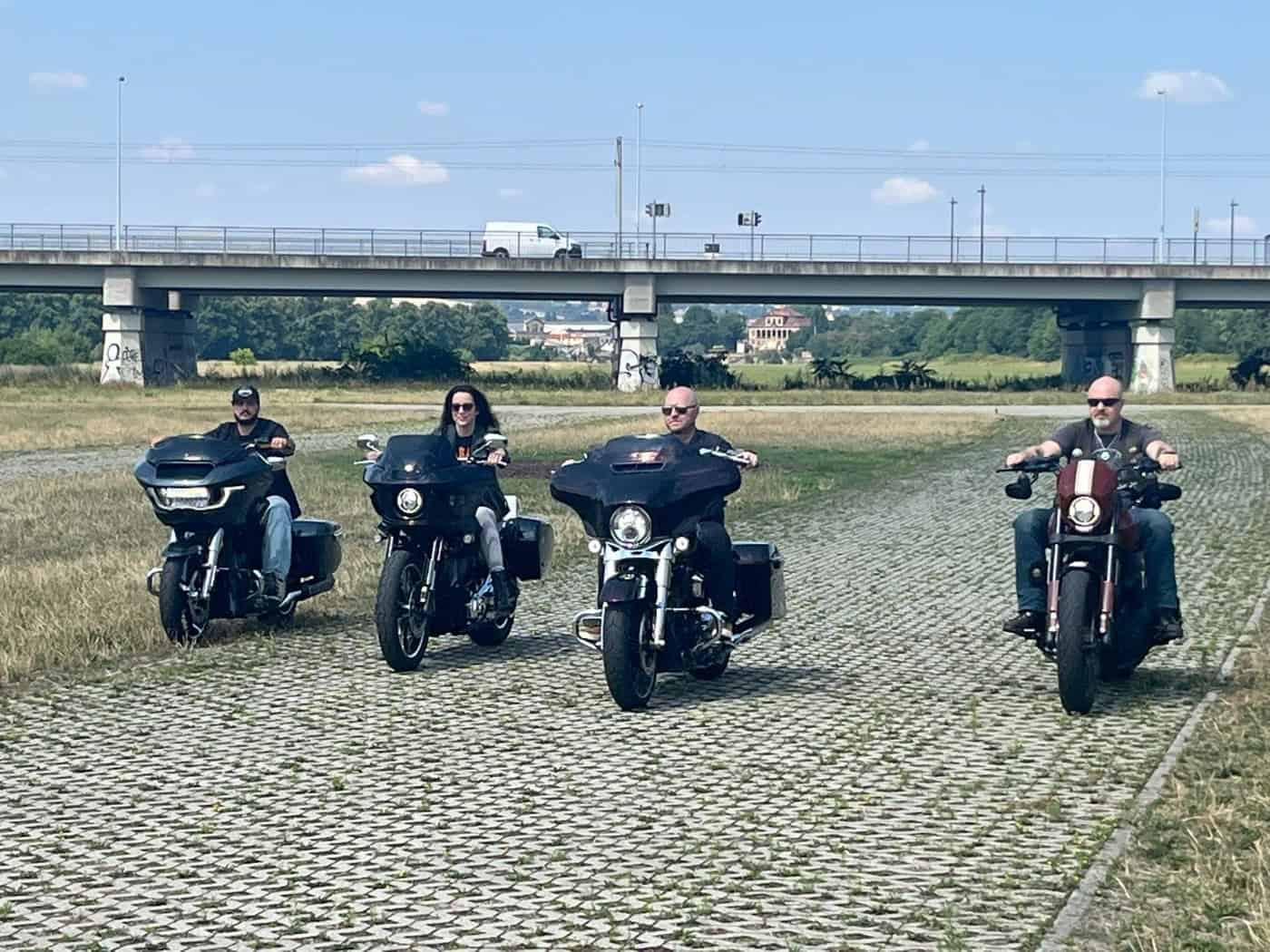 Vier Motorradfahrer auf schweren, dunklen Cruisern fahren nebeneinander auf einem Weg aus Rasengittersteinen durch eine Wiese. Im Hintergrund ist eine große Betonbrücke mit einem weißen Transporter unter leicht bewölktem blauem Himmel zu sehen.