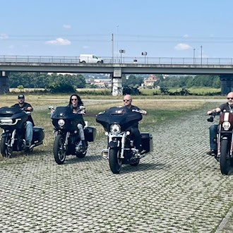 Vier Motorradfahrer auf schweren, dunklen Cruisern fahren nebeneinander auf einem Weg aus Rasengittersteinen durch eine Wiese. Im Hintergrund ist eine große Betonbrücke mit einem weißen Transporter unter leicht bewölktem blauem Himmel zu sehen.