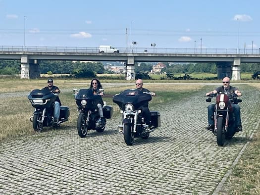 Vier Motorradfahrer auf schweren, dunklen Cruisern fahren nebeneinander auf einem Weg aus Rasengittersteinen durch eine Wiese. Im Hintergrund ist eine große Betonbrücke mit einem weißen Transporter unter leicht bewölktem blauem Himmel zu sehen.