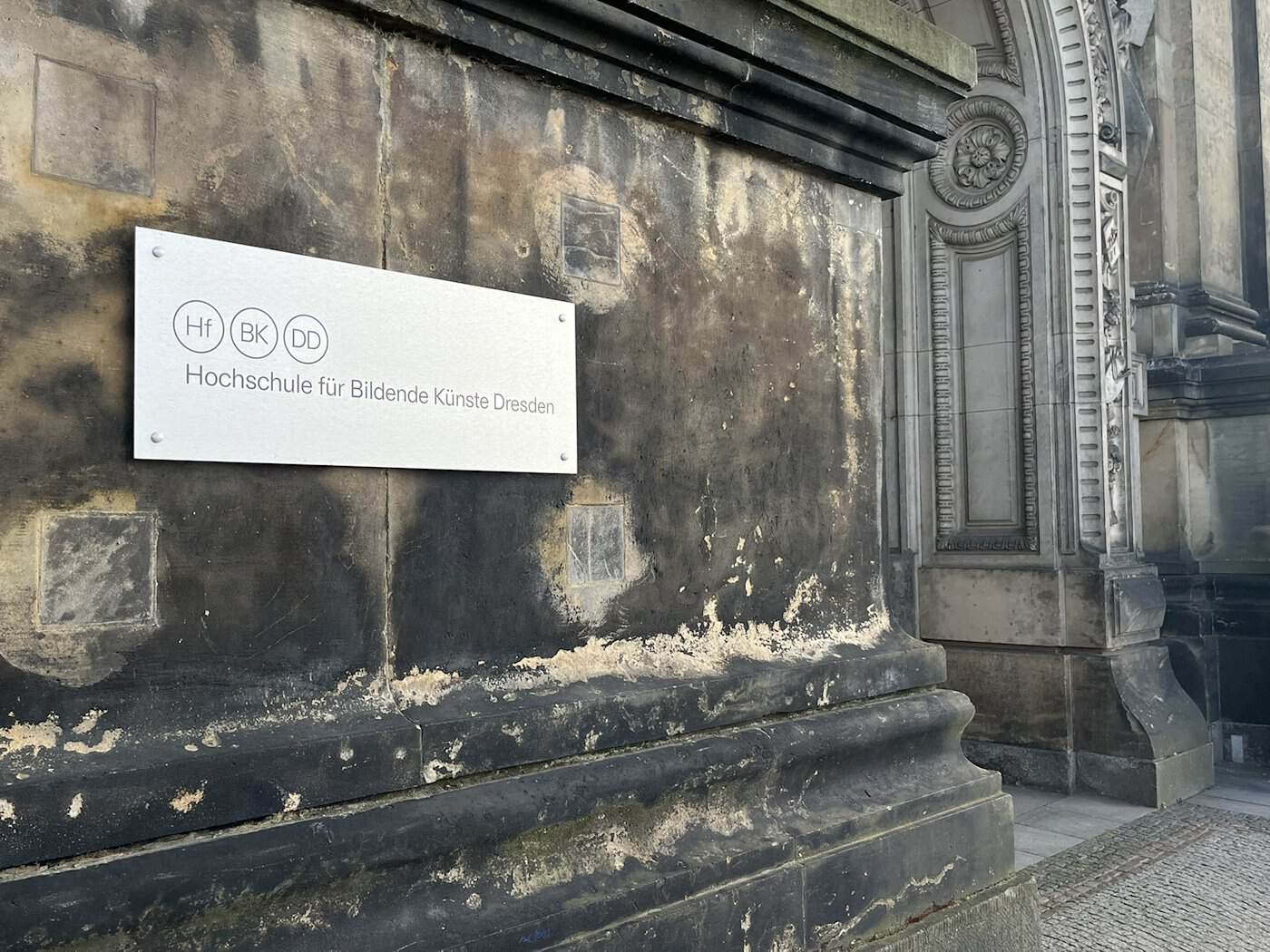 Ein modernes, silbernes Metallschild mit der Aufschrift „Hochschule für Bildende Künste Dresden“ prangt an einer dunkel verwitterten, historischen Sandsteinmauer neben einem barocken Portal.
