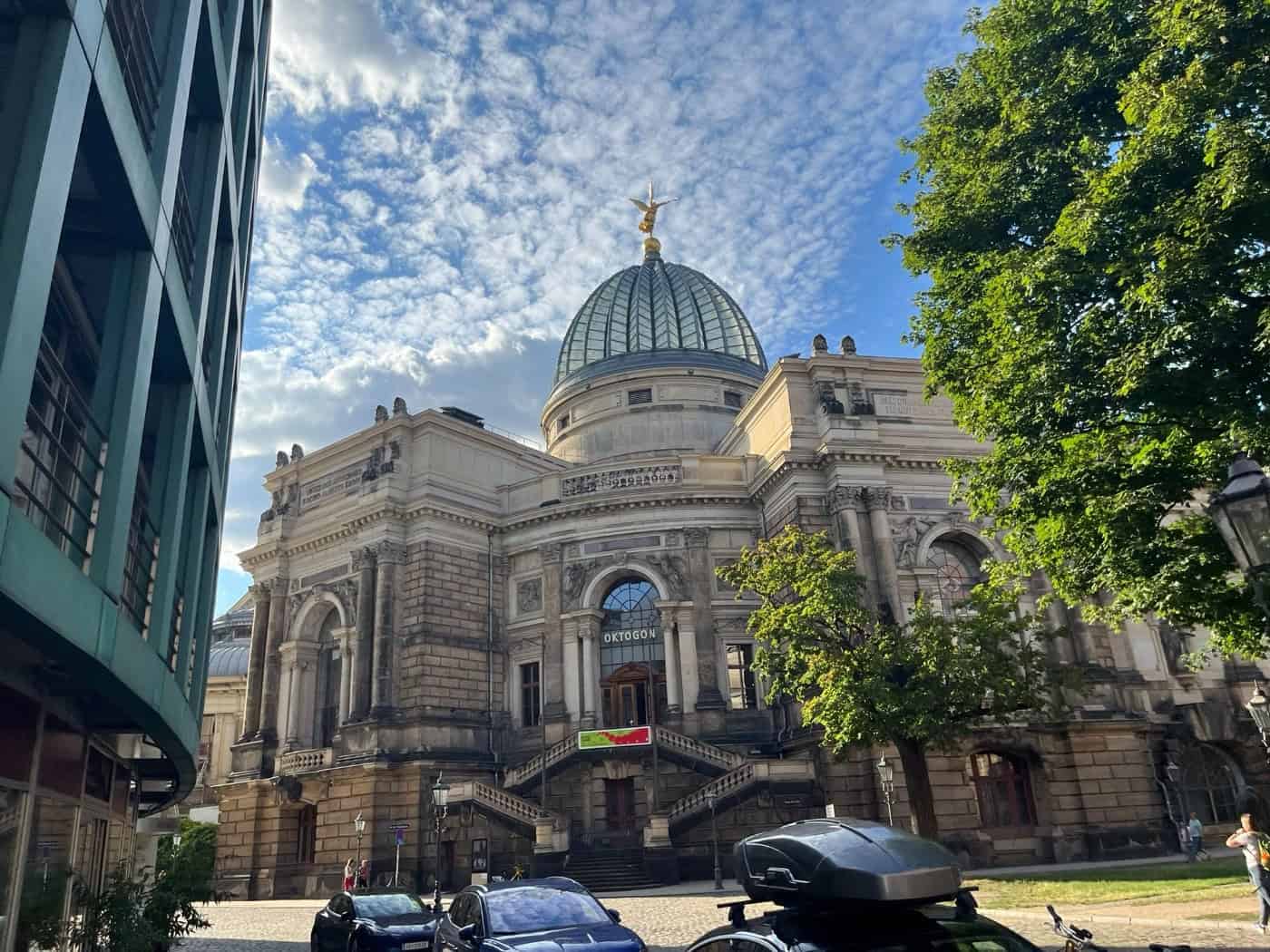 Prachtvolles historisches Akademiegebäude aus Sandstein mit einer großen verzierten Kuppel und goldener Figur an der Spitze. Im Vordergrund stehen parkende Autos, links schneidet die Fassade eines modernen Hauses ins Bild.
