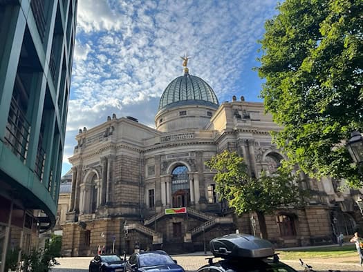 Prachtvolles historisches Akademiegebäude aus Sandstein mit einer großen verzierten Kuppel und goldener Figur an der Spitze. Im Vordergrund stehen parkende Autos, links schneidet die Fassade eines modernen Hauses ins Bild.