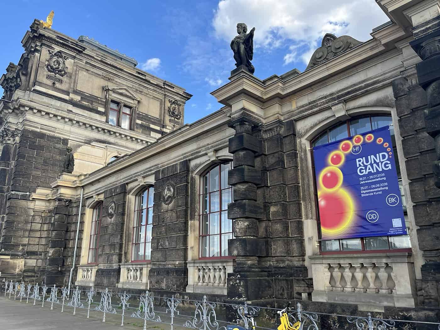 Die Fassade der Kunsthochschule aus dunklem Sandstein unter blauem Himmel. An einem großen Fenster hängt ein blaues Plakat mit bunten Kreisen und der Aufschrift „Rundgang 18.07. - 26.07.2026 Jahresausstellung“.