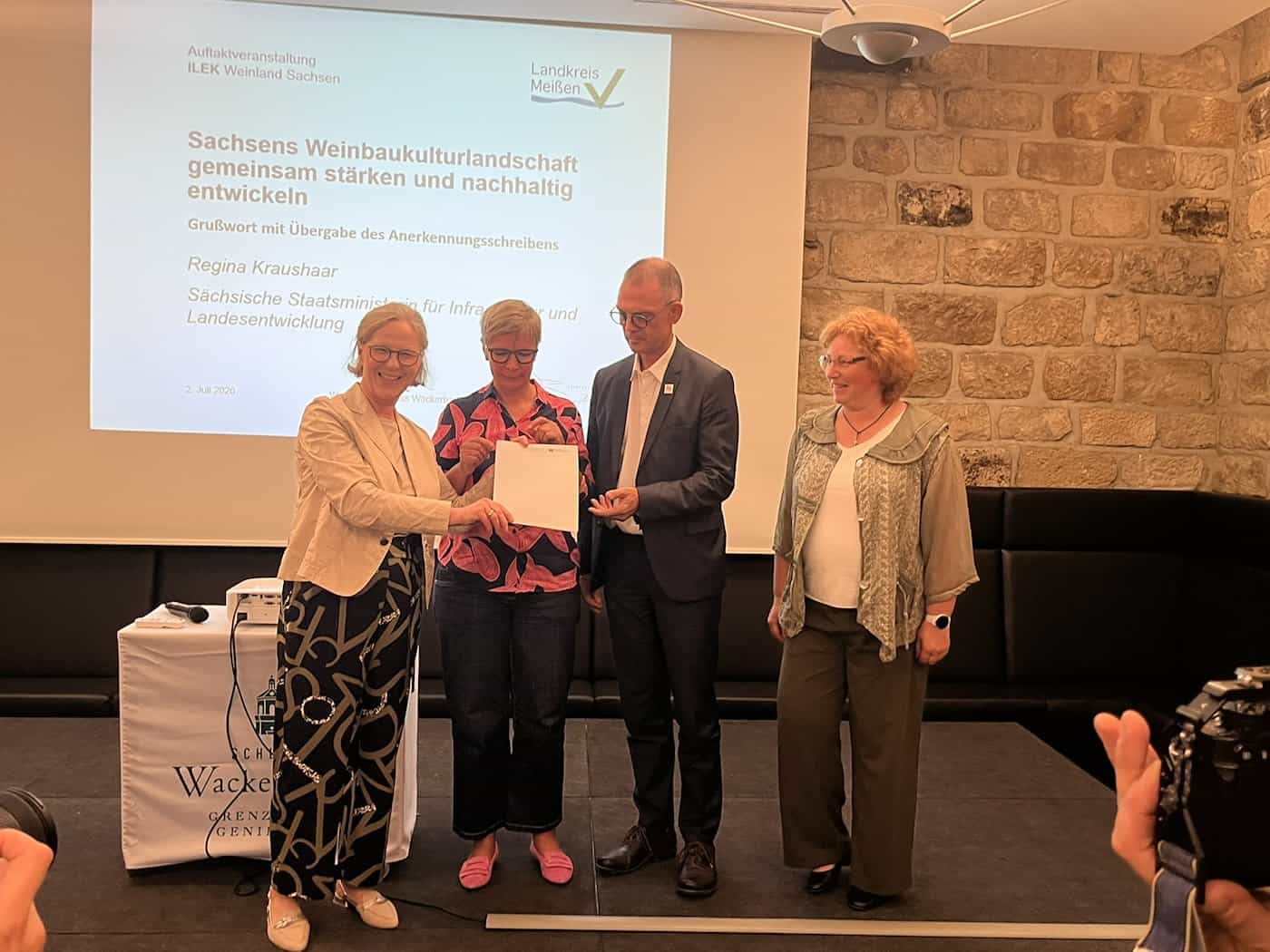 Staatsministerin Regina Kraushaar übergibt lächelnd das ILEK-Anerkennungsschreiben an Dr. Annette Fiss, Landrat Ralf Hänsel und eine weitere Beteiligte auf einer Bühne vor einer Präsentationsleinwand.