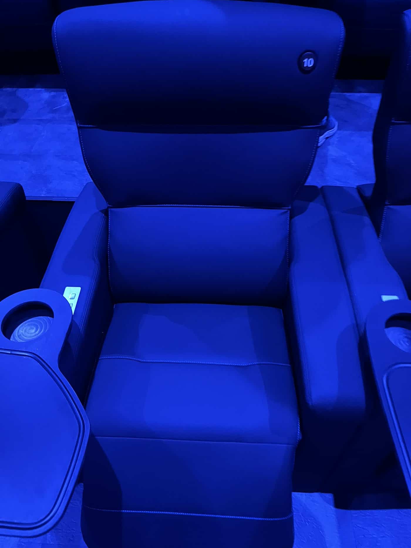 Nahaufnahme eines luxuriösen, dunkelblauen Kinosessels mit verstellbarer Fuß- und Rückenlehne unter blauem Licht. Am Armrest befinden sich silberne Bedienknöpfe sowie ein integrierter Becherhalter. Am Kopfende ist die Sitznummer 10 zu sehen.