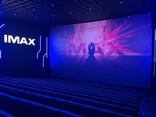 Blick in einen modernen Kinosaal im blauen Licht. Auf einer riesigen, gebogenen Leinwand ist eine Film-Animation zu sehen. Links an der Wand leuchtet ein weißer IMAX-Schriftzug über den ansteigenden Reihen dunkelblauer Sitze.