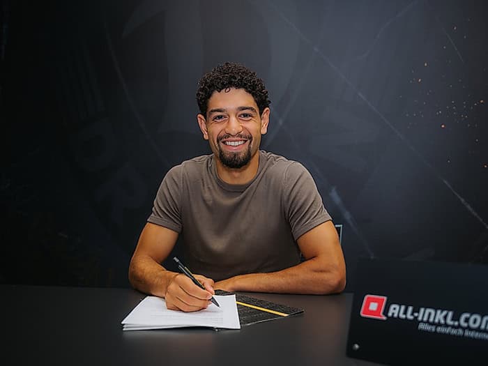 Ben Bobzien sitzt an einem Tisch und unterschreibt seinen Vertrag bei Dynamo Dresden. Vor ihm liegen die Vertragsunterlagen, während er ein schwarzes T-Shirt trägt.