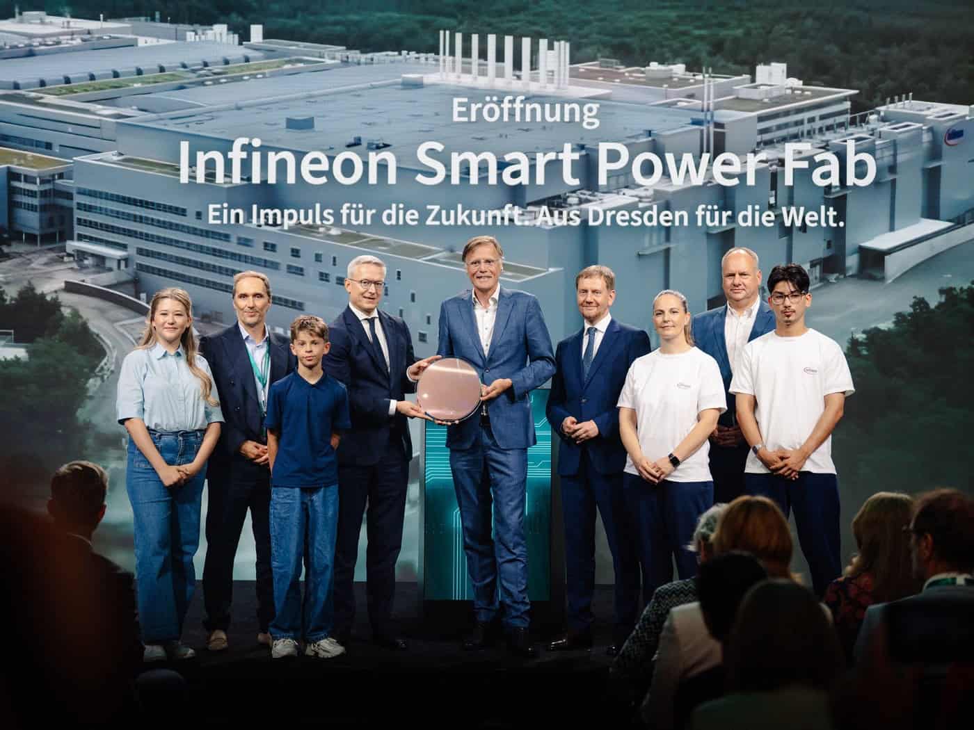 Neun Personen stehen auf einer Bühne vor einer großen Leinwand mit der Aufschrift „Eröffnung Infineon Smart Power Fab“. In der Mitte halten zwei Männer eine runde Halbleiter-Siliziumscheibe (Wafer). Im Vordergrund ist das Publikum von hinten zu sehen.