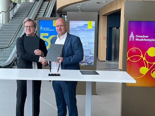 Jan Vogler und Oberbürgermeister Dirk Hilbert stehen lächelnd hinter einem weißen Stehtisch, reichen sich die Hand und halten jeweils eine schwarze Mappe. Im Hintergrund sind digitale Werbetafeln der Dresdner Musikfestspiele zu sehen.