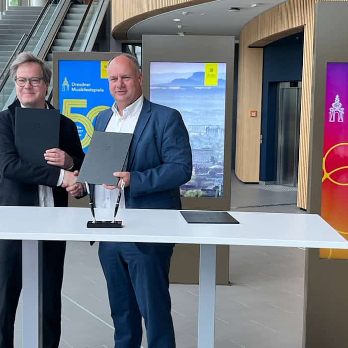 Jan Vogler und Oberbürgermeister Dirk Hilbert stehen lächelnd hinter einem weißen Stehtisch, reichen sich die Hand und halten jeweils eine schwarze Mappe. Im Hintergrund sind digitale Werbetafeln der Dresdner Musikfestspiele zu sehen.