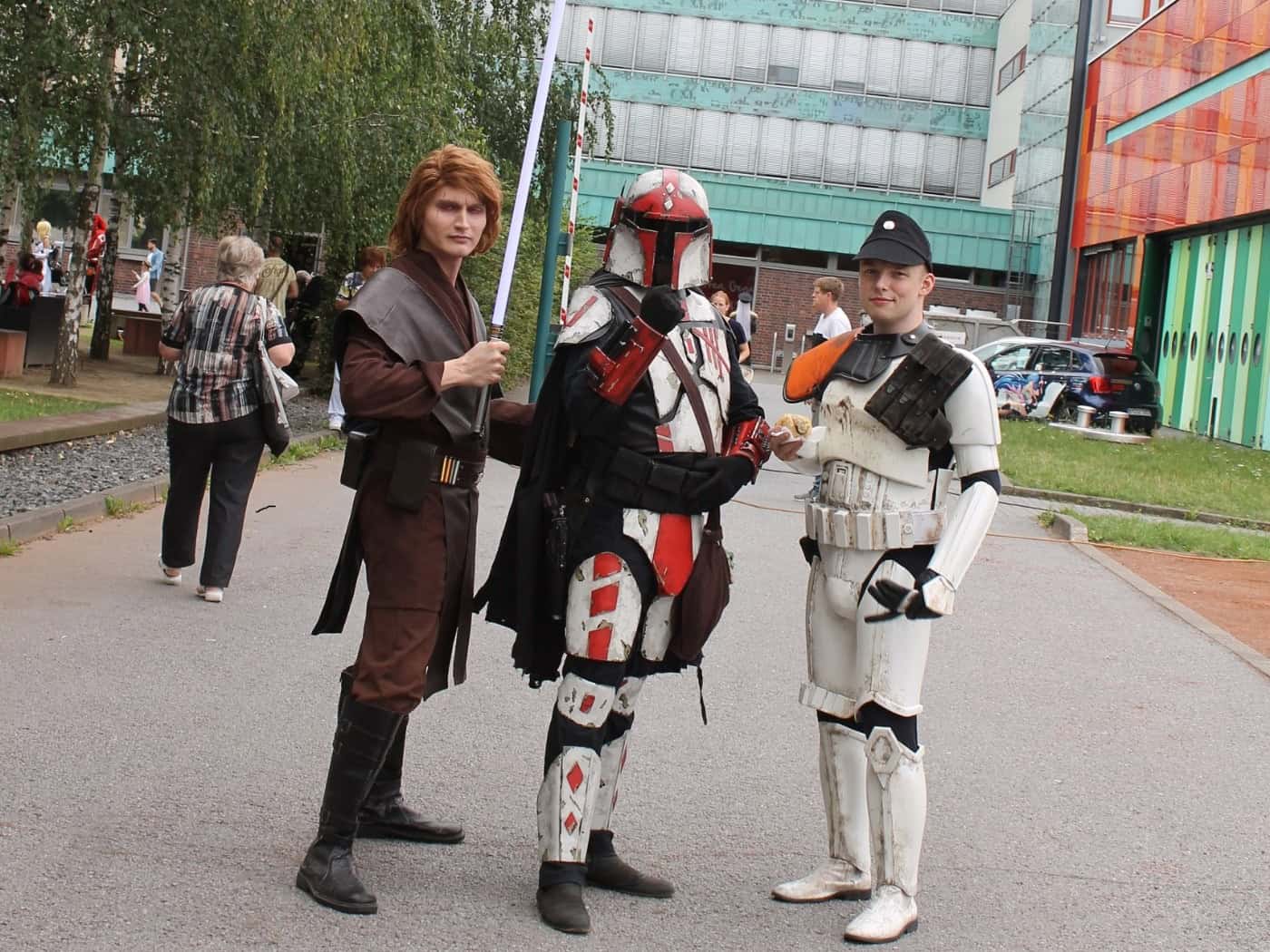 Drei Personen in detaillierten Star-Wars-Kostümen - ein Jedi mit Lichtschwert, ein Mandalorianer und ein Soldat in weißer Rüstung - stehen vor dem hohen, hellblauen Gebäude des Hauses der Presse in Dresden auf einem Asphaltweg. Im Hintergrund wachsen Bäume.