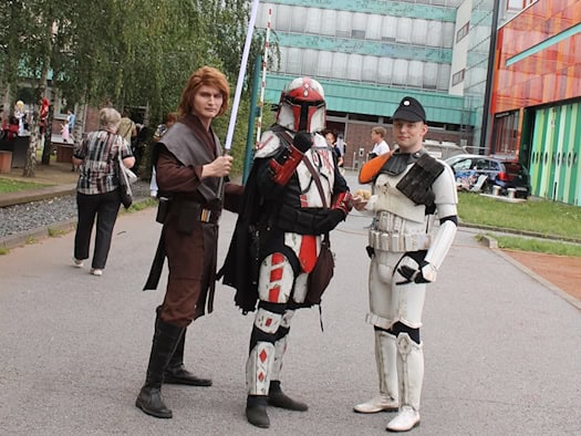 Drei Personen in detaillierten Star-Wars-Kostümen - ein Jedi mit Lichtschwert, ein Mandalorianer und ein Soldat in weißer Rüstung - stehen vor dem hohen, hellblauen Gebäude des Hauses der Presse in Dresden auf einem Asphaltweg. Im Hintergrund wachsen Bäume.