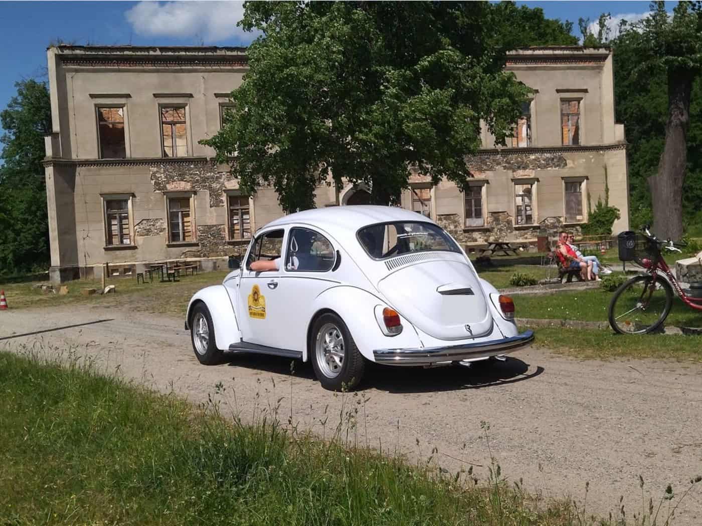 Ein weißer VW-Käfer fährt über einen Sandweg. Im Hintergrund ein stark verfallenes Herrenhaus bzw. Schloss und ein Garten, in dem mehrere Personen auf einer Bank sitzen.