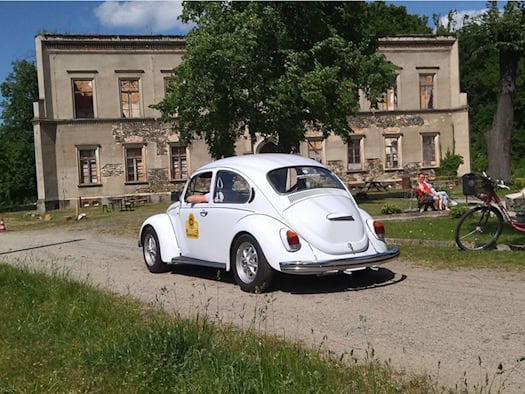 Ein weißer VW-Käfer fährt über einen Sandweg. Im Hintergrund ein stark verfallenes Herrenhaus bzw. Schloss und ein Garten, in dem mehrere Personen auf einer Bank sitzen.