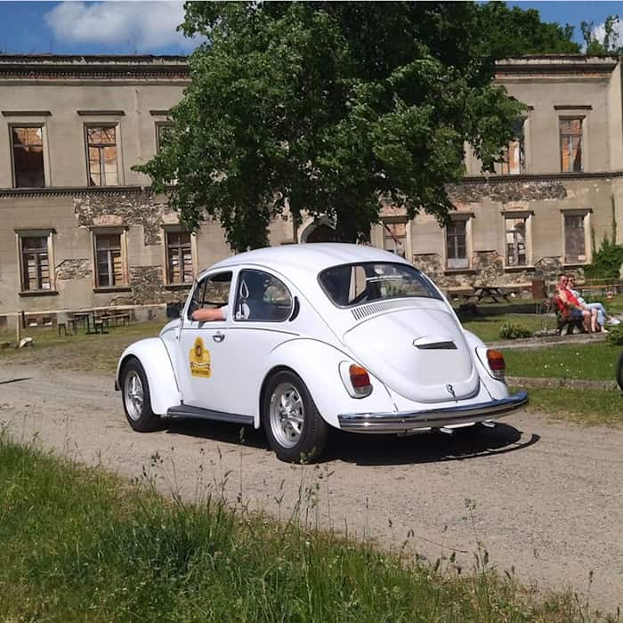 Ein weißer VW-Käfer fährt über einen Sandweg. Im Hintergrund ein stark verfallenes Herrenhaus bzw. Schloss und ein Garten, in dem mehrere Personen auf einer Bank sitzen.