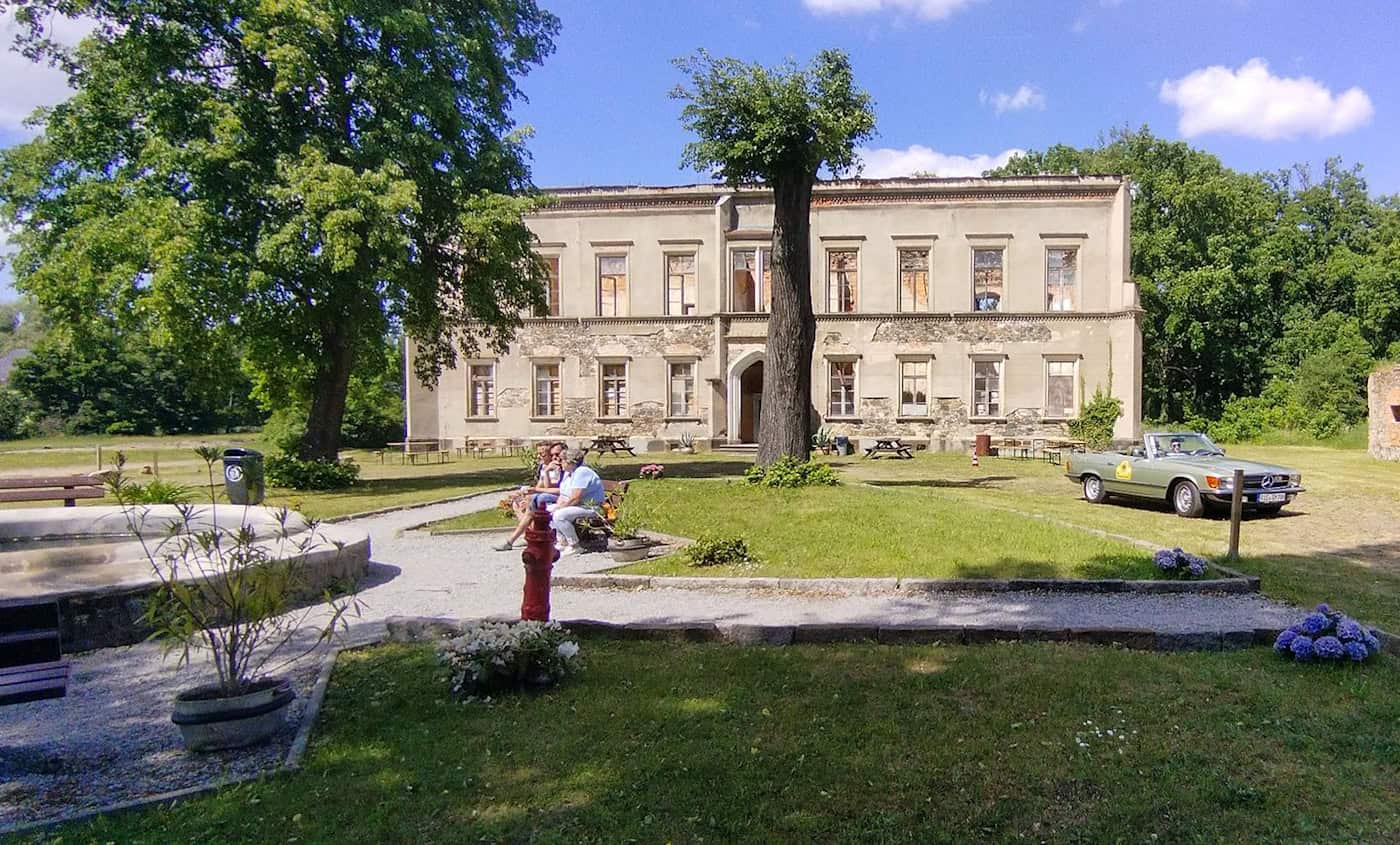 Blick durch einen Park auf ein zweistöckiges Herrenhaus bzw. Schloss. Links ein steinernes Brunnenbecken, in der Mitte drei Personen sitzend auf einer Bank, rechts im Bild ein Mercedes-Cabrio-Oldtimer.
