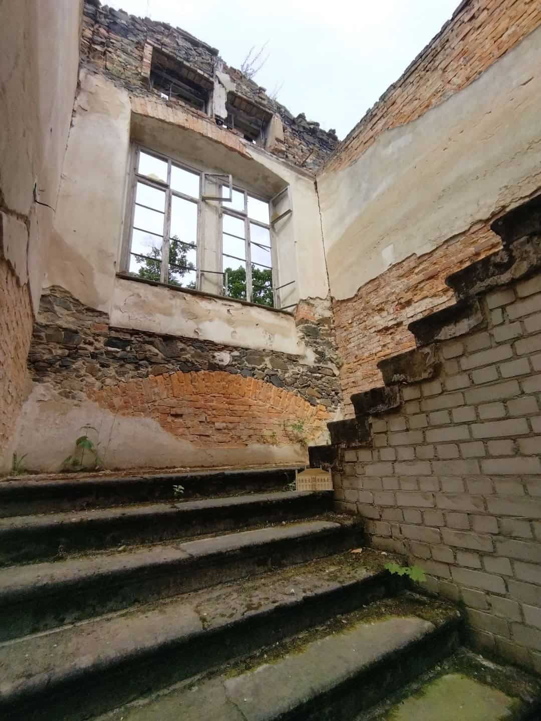 Blick in ein stark verfallenes Treppenhaus. Die Decke fehlt völlig, die Wände sind rissig, bröckeln und sind weitgehend unverputzt.