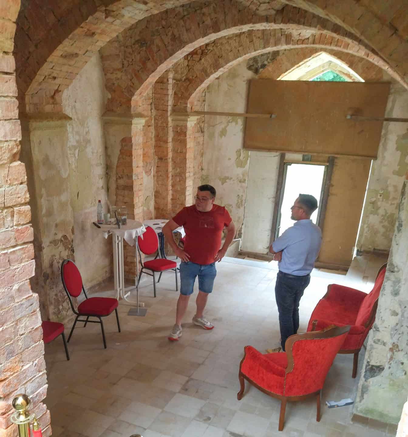 Blick in den Eingangsbereiches eines Hauses, in dem zwei Männer stehen und sich unterhalten. Die Wände sind unverputzt, teils beschädigt. Im Raum stehen zwei rote Sessel, ein paar Stühle und ein Stehtisch mit Infomaterial.