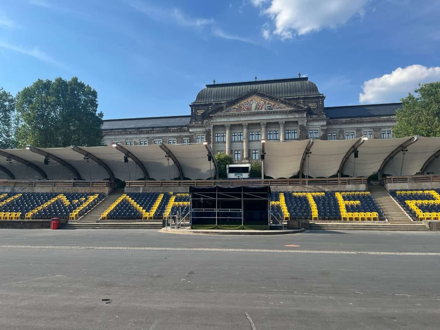 Leere Zuschauerränge einer Open-Air-Bühne mit blauen und gelben Sitzen, die das Wort „FILMNÄCHTE“ bilden. Dahinter steht ein großes historisches Regierungsgebäude mit einer verzierten Fassade unter blauem Himmel mit weißen Wolken.