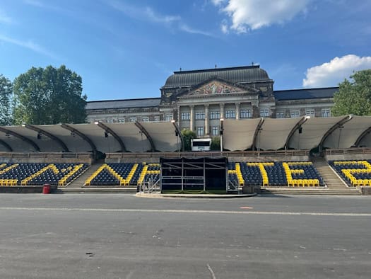 Leere Zuschauerränge einer Open-Air-Bühne mit blauen und gelben Sitzen, die das Wort „FILMNÄCHTE“ bilden. Dahinter steht ein großes historisches Regierungsgebäude mit einer verzierten Fassade unter blauem Himmel mit weißen Wolken.