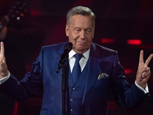 Nahaufnahme von Roland Kaiser im eleganten, blauen dreiteiligen Anzug mit Krawatte hinter einem Mikrofon. Er blickt lächelnd nach vorn und hebt beide Hände mit ausgestreckten Fingern zum Gruß. Der Hintergrund ist dunkel mit dezenten roten Lichteffekten.