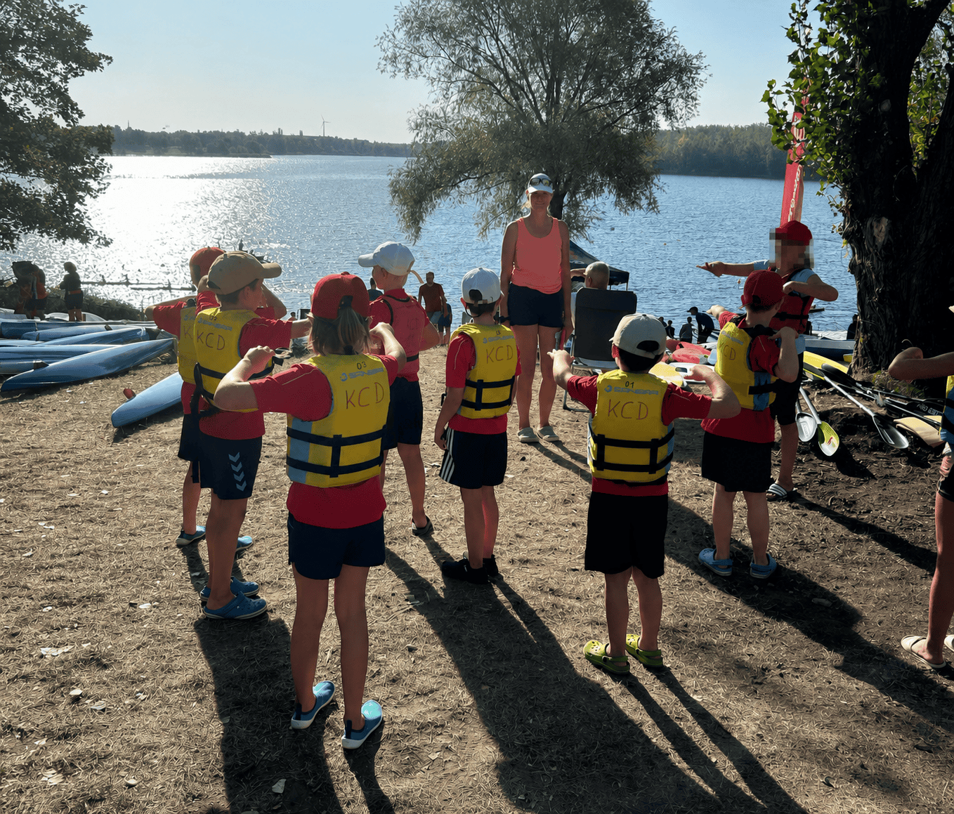 Jugendgruppe des Kanu Clubs Dresden mit Trainerin Anja Souček am Ufer Eine Gruppe von Kindern in roten Shirts und gelben Schwimmwesten mit der Aufschrift KCD steht im Kreis am sandigen Ufer eines Sees. Vor ihnen steht eine Frau im ärmellosen Shirt im hellen Gegenlicht der Sonne.