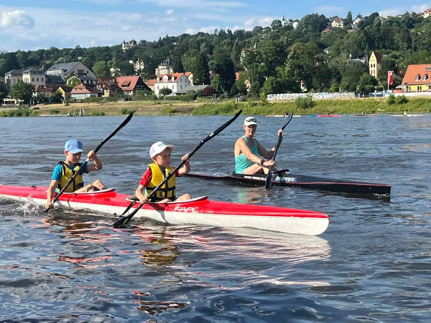 Zwei Kinder in einem rot-weißen Kajak paddeln auf einem breiten Fluss, gefolgt von einer Frau in einem schwarzen Boot. Im Hintergrund erstreckt sich ein grüner Hang mit historischen Häusern unter leicht bewölktem blauen Himmel.