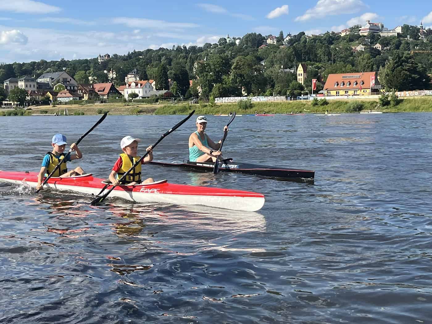 Zwei Kinder in einem rot-weißen Kajak paddeln auf einem breiten Fluss, gefolgt von einer Frau in einem schwarzen Boot. Im Hintergrund erstreckt sich ein grüner Hang mit historischen Häusern unter leicht bewölktem blauen Himmel.