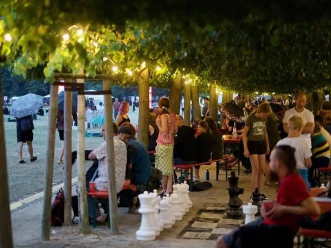 Menschen sitzen am Abend an Biertischen unter einer Allee aus beleuchteten Bäumen. Im Vordergrund sind große, weiße und schwarze Schachfiguren auf einem XXL-Spielfeld zu sehen. Links erstreckt sich eine Wiese, auf der eine Person mit Regenschirm läuft.