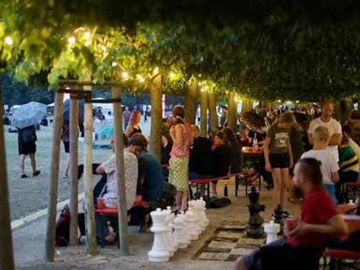 Menschen sitzen am Abend an Biertischen unter einer Allee aus beleuchteten Bäumen. Im Vordergrund sind große, weiße und schwarze Schachfiguren auf einem XXL-Spielfeld zu sehen. Links erstreckt sich eine Wiese, auf der eine Person mit Regenschirm läuft.