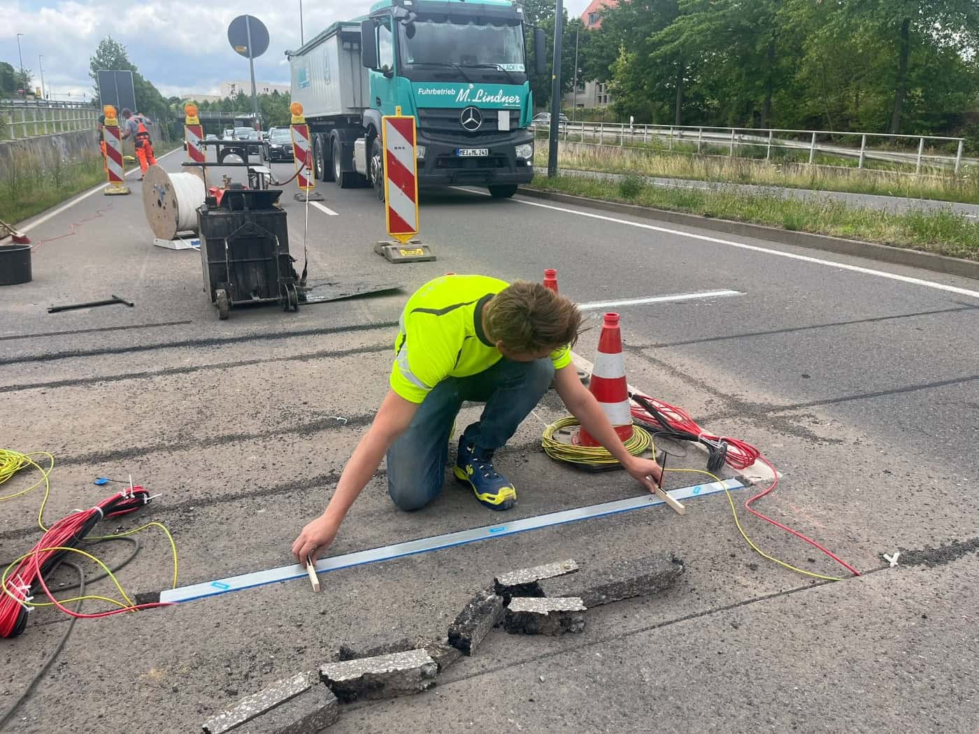 Ein Techniker im neongelben T-Shirt und blauer Jeans kniet auf der Straße und setzt eine lange Kistler-Metallschiene präzise in eine Fräsrinne im Asphalt ein. Im Hintergrund steht ein großer grüner Lkw.