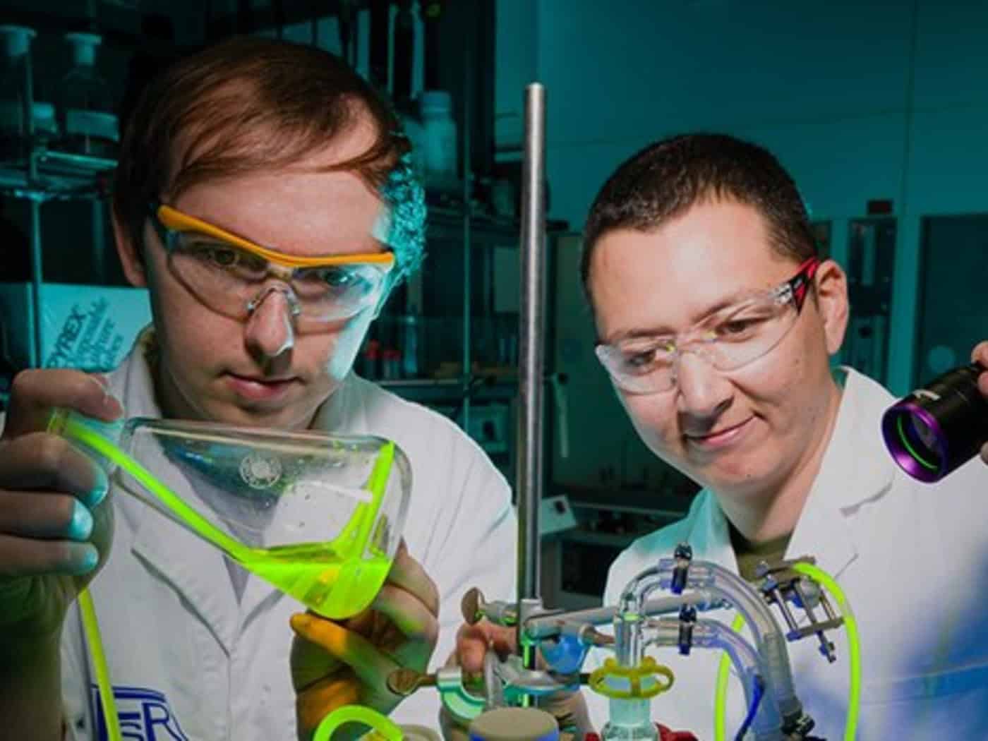 Zwei Forscher in Laborzeugen und Schutzbrillen arbeiten in einem abgedunkelten Raum. Der linke hält einen Kolben mit neongrün leuchtender Flüssigkeit. Der rechte zielt mit einer UV-Taschenlampe auf ein gläsernes Reaktorsystem mit grünen Schläuchen.