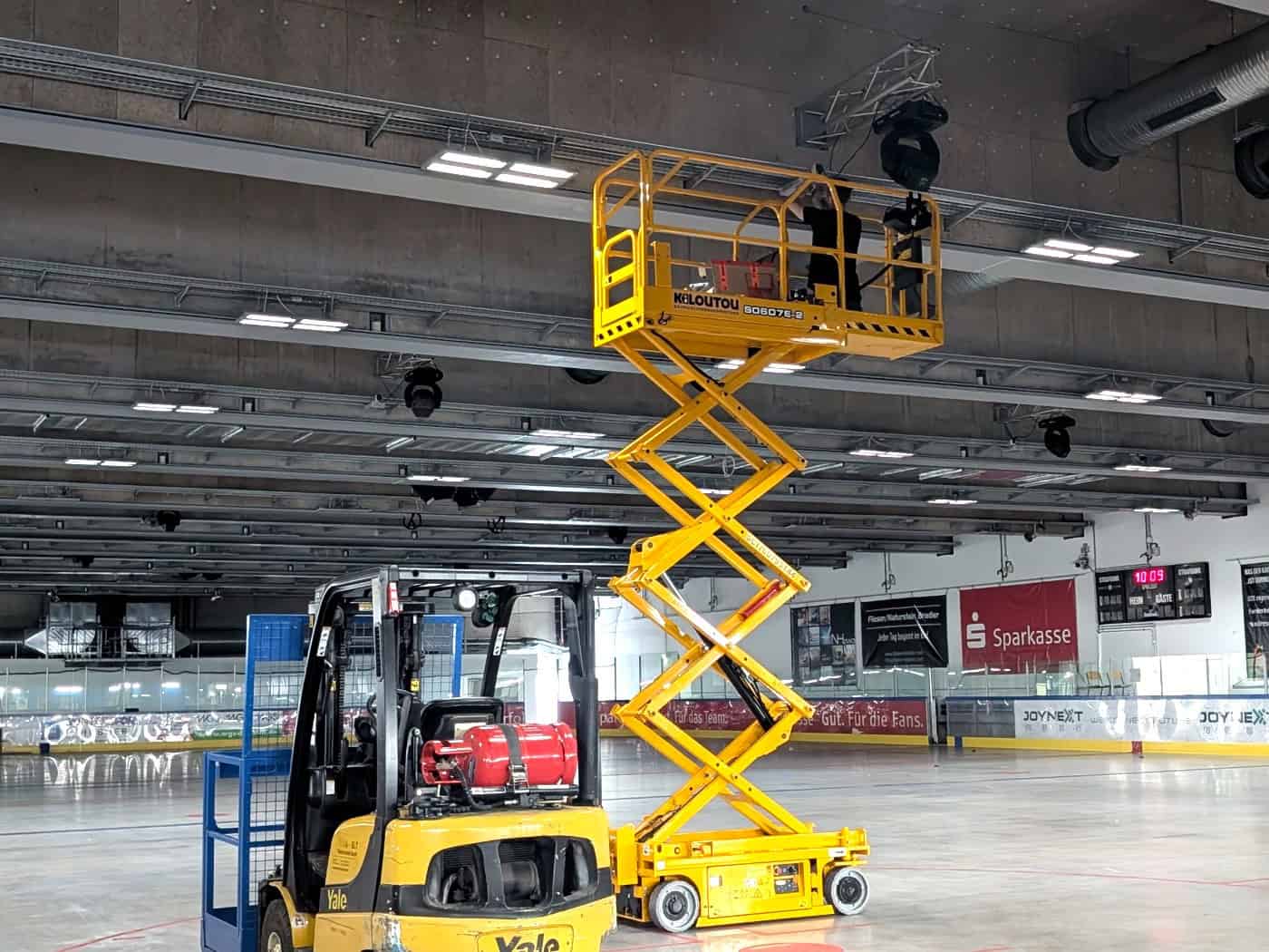 Eine große, eisfreie Eishalle mit grauem Betonboden. Im Zentrum steht eine hoch ausgefahrene gelbe Scherenbühne mit einem Handwerker an der Decke. Im Vordergrund parkt ein gelber Gabelstapler. An der Decke leuchten bereits neue, quadratische LED-Panels.