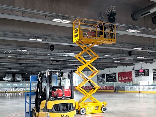 Eine große, eisfreie Eishalle mit grauem Betonboden. Im Zentrum steht eine hoch ausgefahrene gelbe Scherenbühne mit einem Handwerker an der Decke. Im Vordergrund parkt ein gelber Gabelstapler. An der Decke leuchten bereits neue, quadratische LED-Panels.