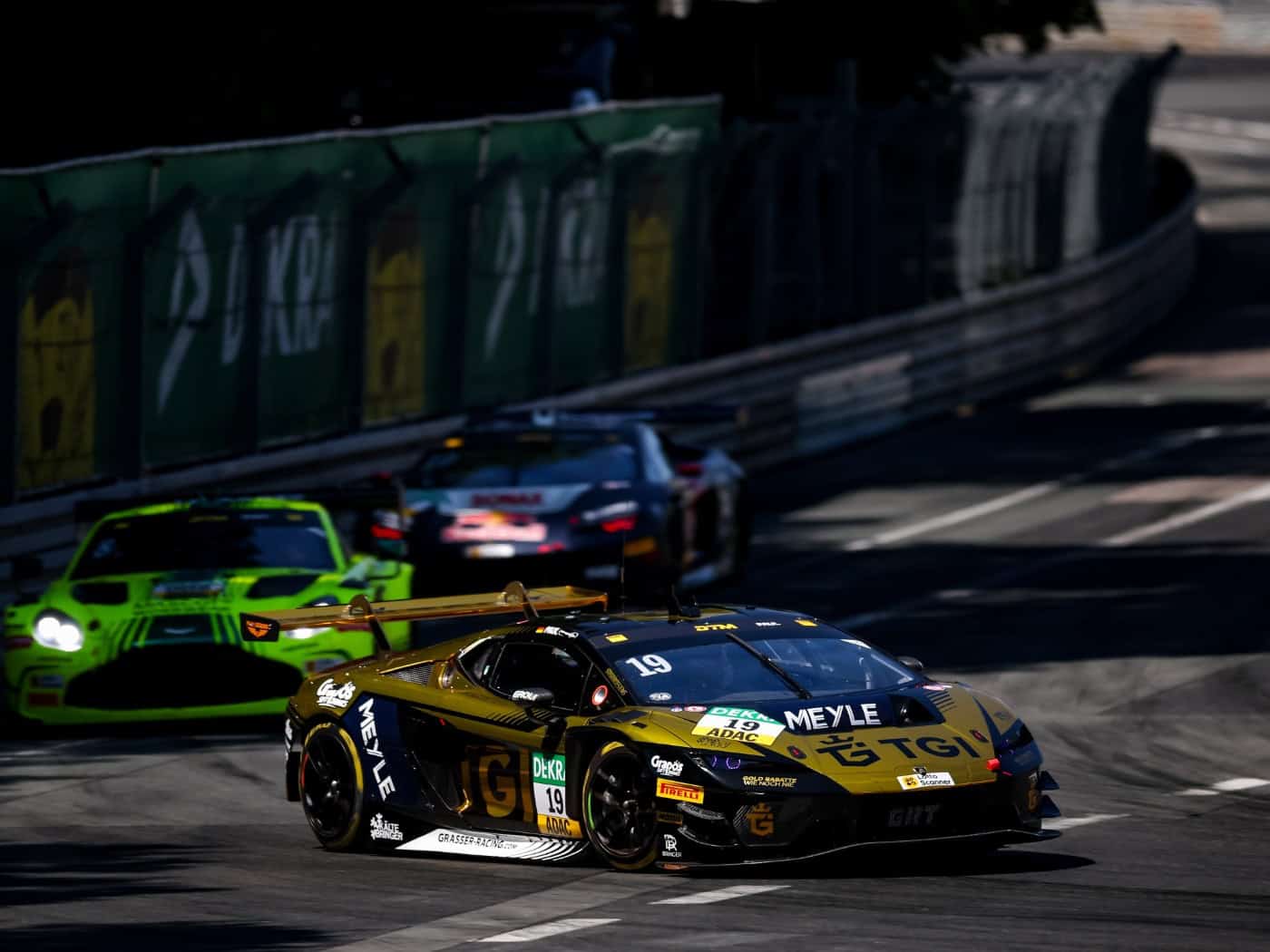 Ein gold-schwarzer Lamborghini-Rennwagen mit der Nummer 19 fährt in Führung liegend auf einem engen Stadtkurs. Im Hintergrund folgen weitere Rennautos, darunter ein grüner Aston Martin. Hohe Leitplanken säumen die sonnenbeschienene Rennstrecke.