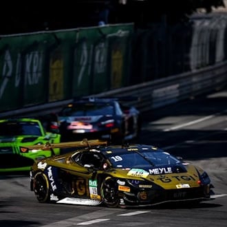 Ein gold-schwarzer Lamborghini-Rennwagen mit der Nummer 19 fährt in Führung liegend auf einem engen Stadtkurs. Im Hintergrund folgen weitere Rennautos, darunter ein grüner Aston Martin. Hohe Leitplanken säumen die sonnenbeschienene Rennstrecke.