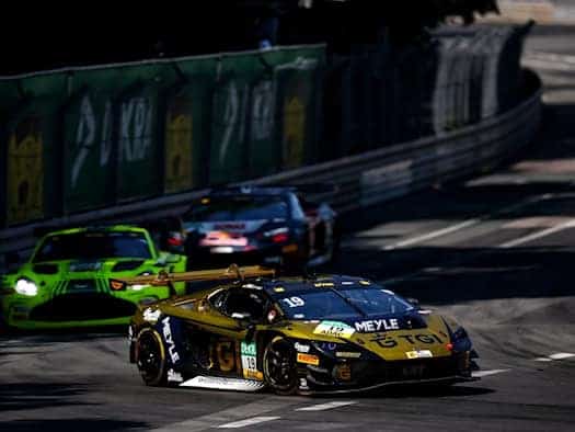 Ein gold-schwarzer Lamborghini-Rennwagen mit der Nummer 19 fährt in Führung liegend auf einem engen Stadtkurs. Im Hintergrund folgen weitere Rennautos, darunter ein grüner Aston Martin. Hohe Leitplanken säumen die sonnenbeschienene Rennstrecke.