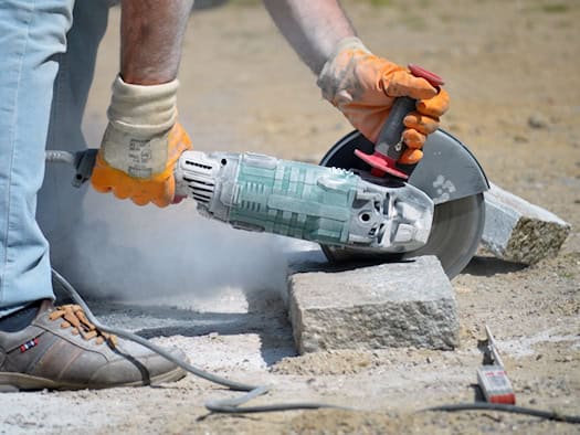 Nahaufnahme von Beinen und Händen eines Handwerkers, der mit einem grünen Winkelschleifer einen grauen Granitstein schneidet. Er trägt orangefarbene Schutzhandschuhe und braune Arbeitsschuhe. Feiner, heller Steinstaub wirbelt in der Luft auf.